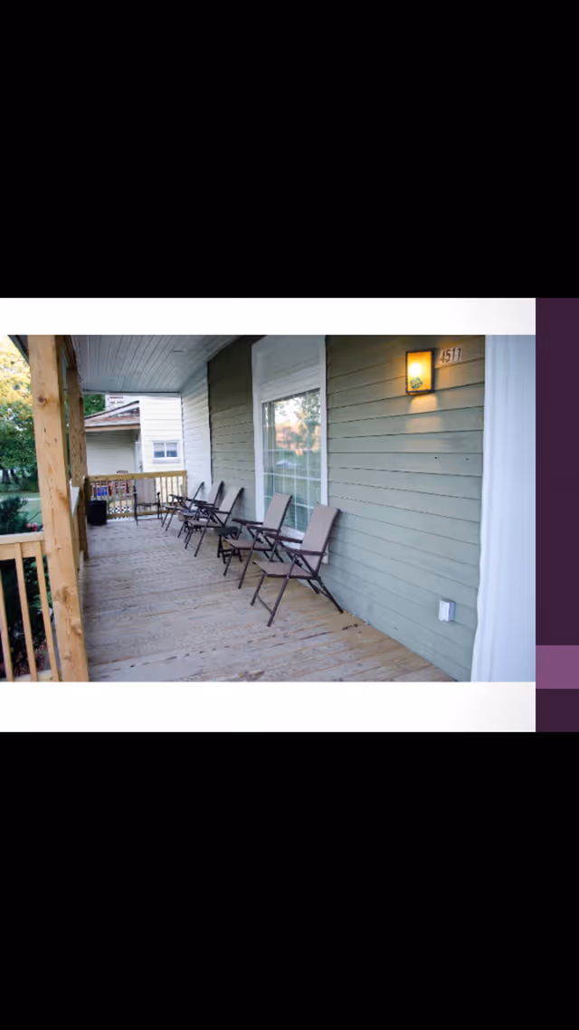 A covered wooden porch with several folding chairs lined up against the exterior wall of a building. The porch has a railing and a light fixture mounted on the wall next to a window. The house number 4511 is visible near the light.