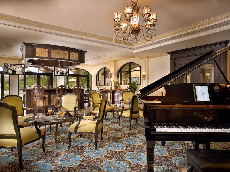Elegant interior room with a grand piano on the right, several yellow upholstered chairs around small round tables, a bar area with high chairs in the background, and large arched windows letting in natural light. The room features a decorative chandelier and a patterned carpet with blue and brown tones.