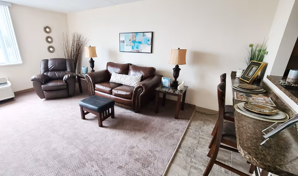 A cozy living room area with a brown leather loveseat and matching armchair, a small black ottoman, two side tables with lamps, and a framed abstract painting on the wall. To the right, there is a kitchen counter with bar stools and place settings. The room has carpeted flooring transitioning to tiled flooring near the kitchen counter.
