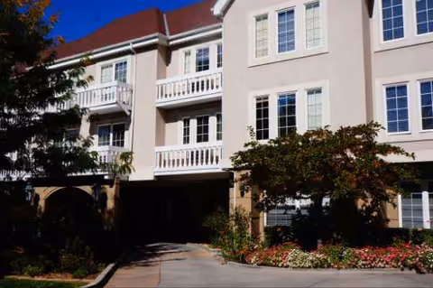 A multi-story senior living building with white balconies, windows, and a landscaped entrance.