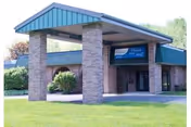 Exterior view of a single-story brick building with a covered entrance supported by brick pillars and a blue roof. There is a sign above the entrance that reads 'Embassy of Tunkhannock'. The building is surrounded by green grass and some trees in the background.