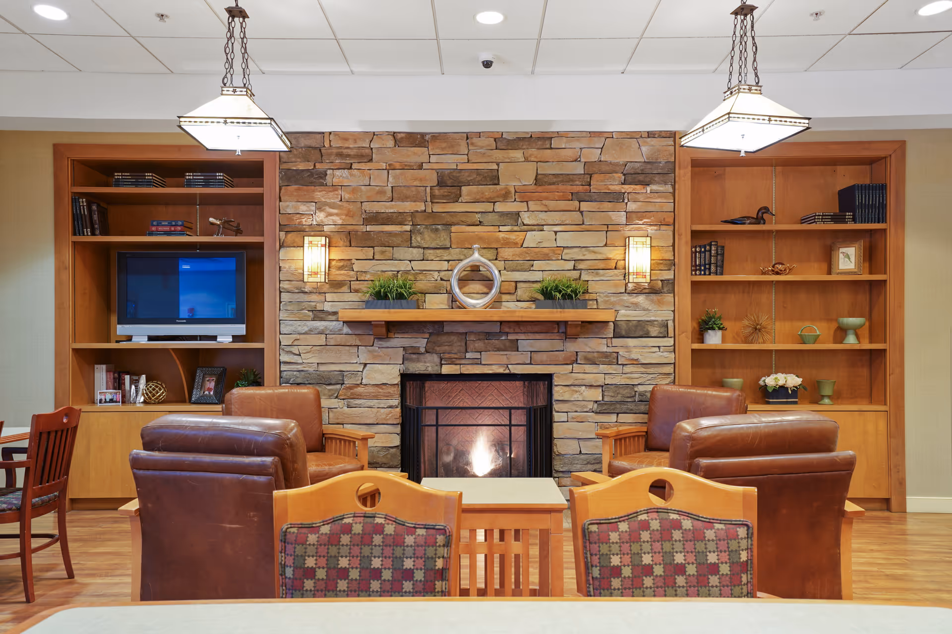Cozy living room area with a stone fireplace in the center, flanked by wooden built-in shelves filled with books and decorative items. Four brown leather armchairs are arranged around a small wooden table in front of the fireplace. Two pendant lights hang from the ceiling, illuminating the space.