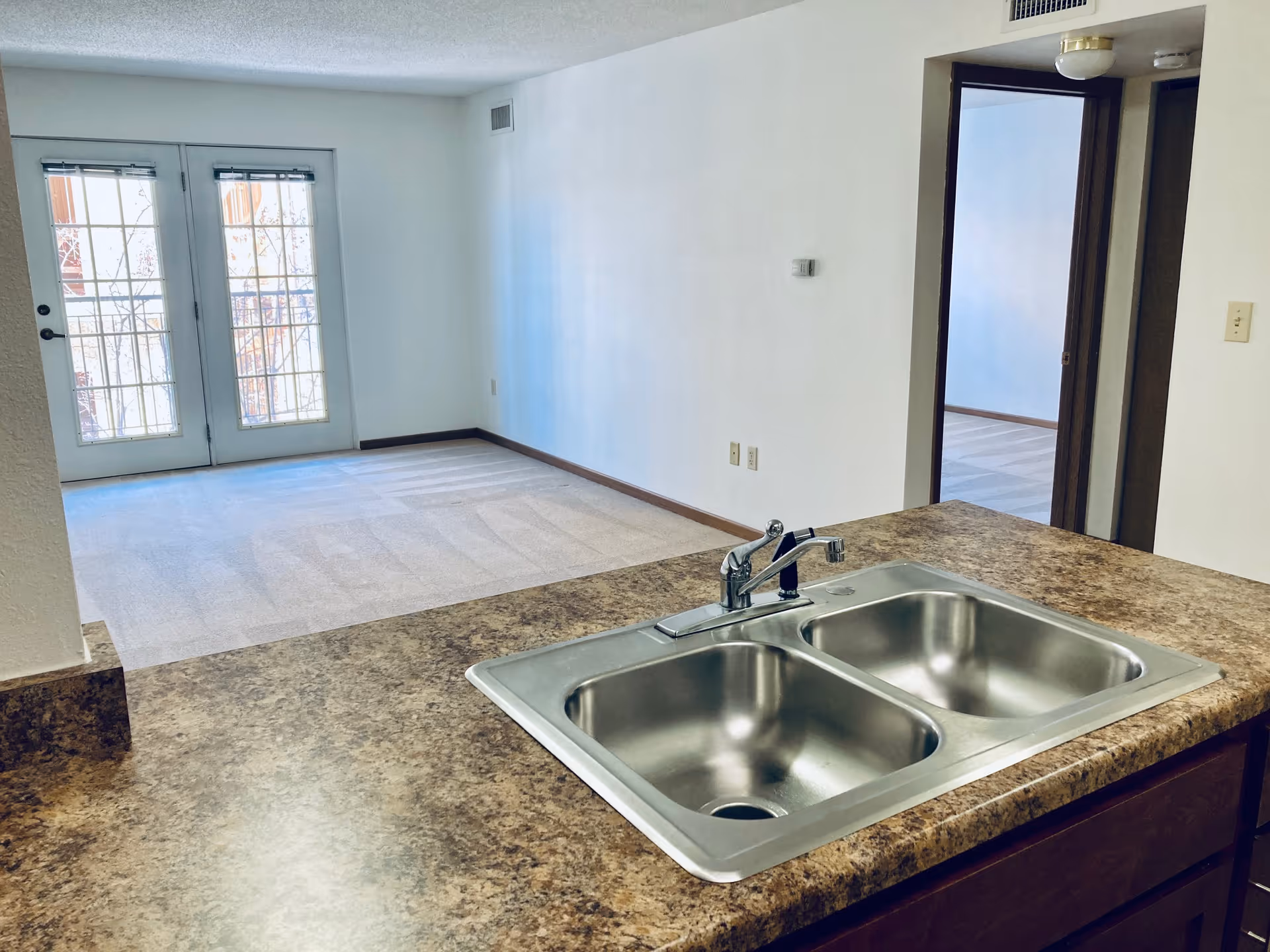 View from a kitchen counter with a double stainless steel sink looking out into a carpeted living area with double glass doors leading to a balcony. There is a doorway on the right side leading to another room.