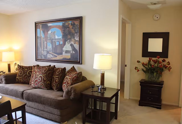 A cozy living room with a brown sofa adorned with patterned cushions, two side tables each holding a lamp, a framed painting of an ancient ruin on the wall above the sofa, and a small dark wooden cabinet with a vase of red flowers and a square mirror above it.