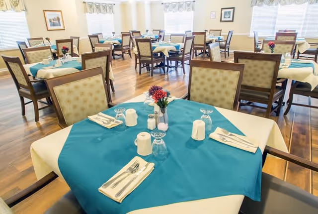 A bright dining room with multiple tables covered in white and teal tablecloths, each set with utensils, napkins, salt and pepper shakers, and a small flower arrangement. The room has wooden floors, cushioned chairs, and windows with valances letting in natural light.