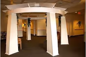 Interior view of a common area in an assisted living facility featuring a large white pergola-like structure with four thick pillars. The room has beige walls, carpeted floors, and some chairs and plants visible in the background.