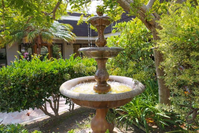 A three-tiered stone fountain surrounded by lush greenery and trees in a garden courtyard by a building.