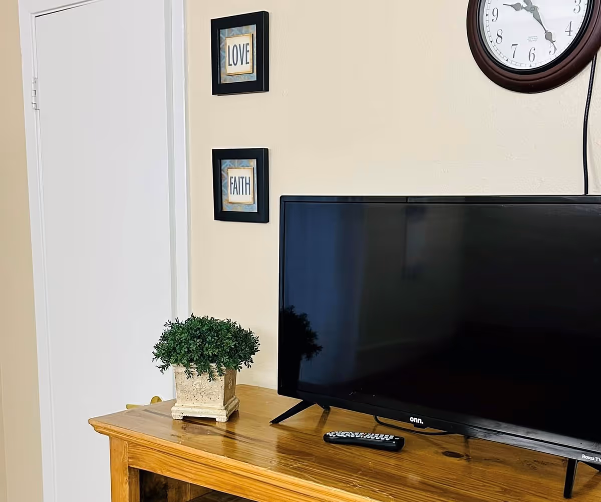 A wooden TV stand with a flat-screen TV and a remote control on top. Next to the TV is a small potted plant in a square stone-like pot. On the wall above the TV are two framed pictures with the words 'LOVE' and 'FAITH'. A round wall clock is partially visible on the upper right side of the image.
