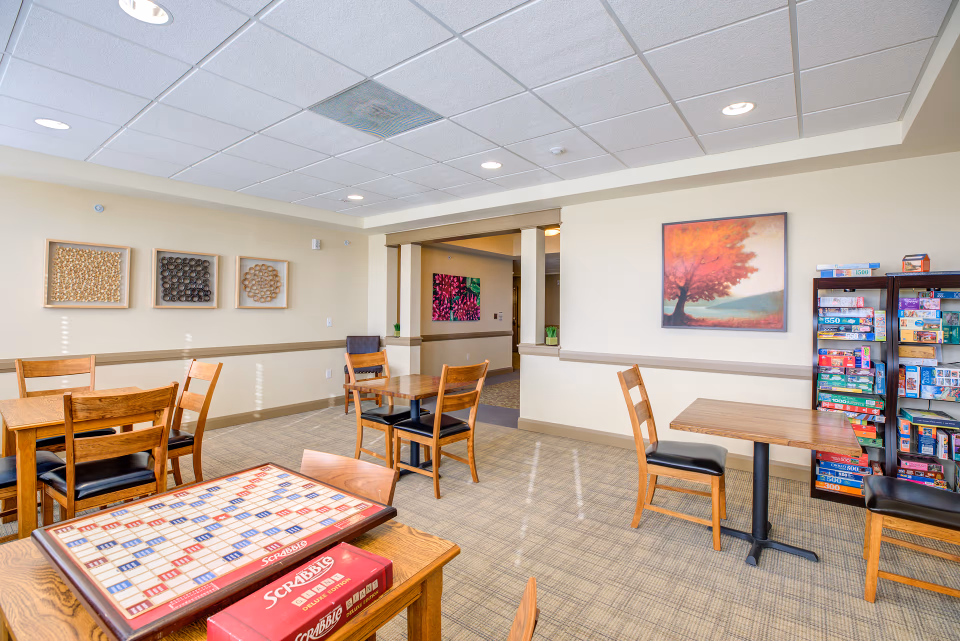A bright and spacious game room with several wooden tables and chairs. One table in the foreground has a Scrabble board game set up. Shelves filled with various board games are visible against the wall on the right. The walls are decorated with framed artwork, including a painting of a tree with red leaves and three abstract circular designs. The room has a beige carpet and a white drop ceiling with recessed lighting.