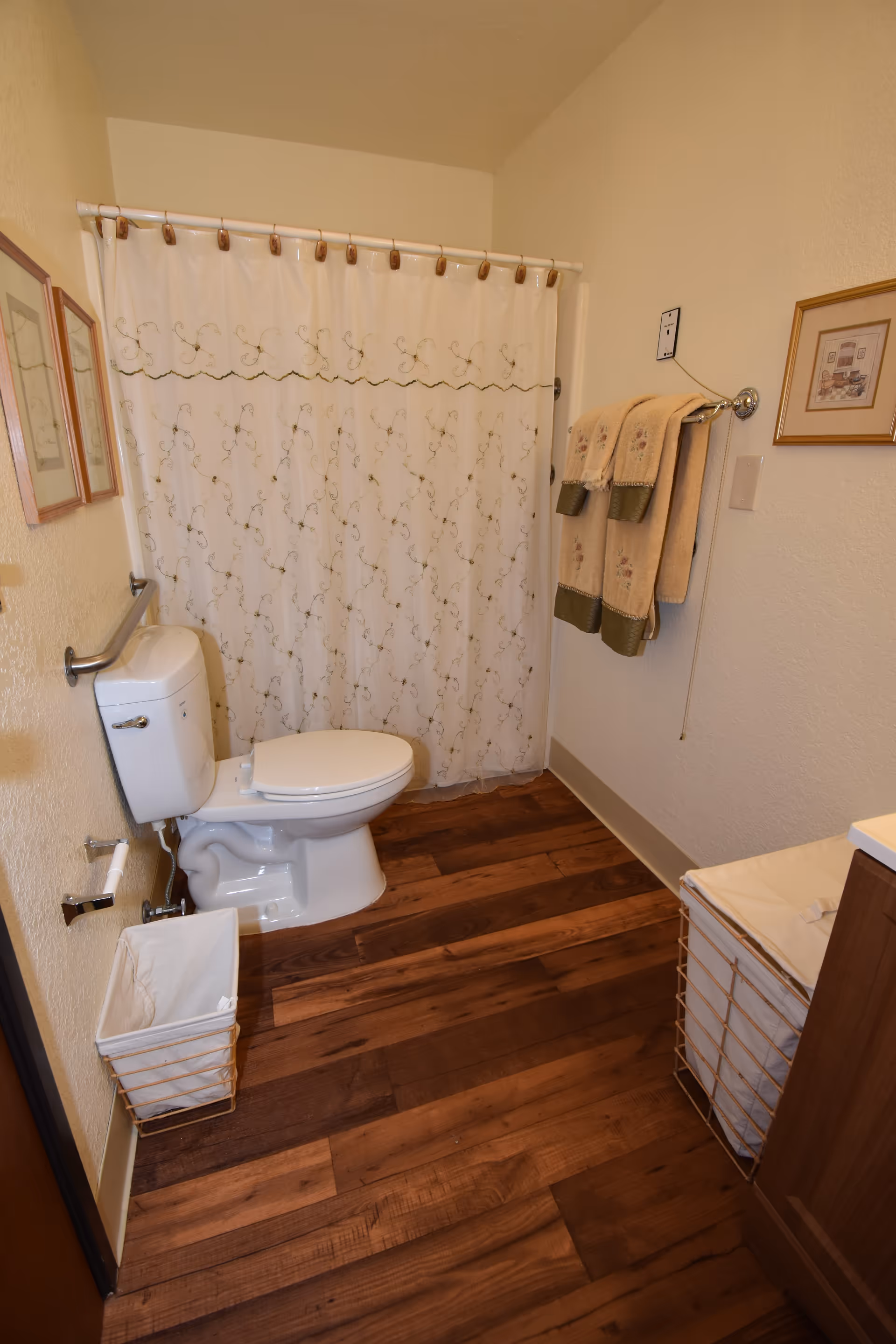 A bathroom with a white toilet, a shower with a white curtain featuring a floral pattern, wooden flooring, a towel rack with beige and green towels, a wastebasket, and framed pictures on the walls.