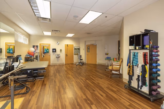 A spacious therapy or exercise room with wooden flooring, a treatment table, parallel bars, exercise balls, and a rack holding various weights and resistance bands. The room has beige walls, a large mirror on one side, and several wooden doors. There is also a chair and a small sink on the far wall.