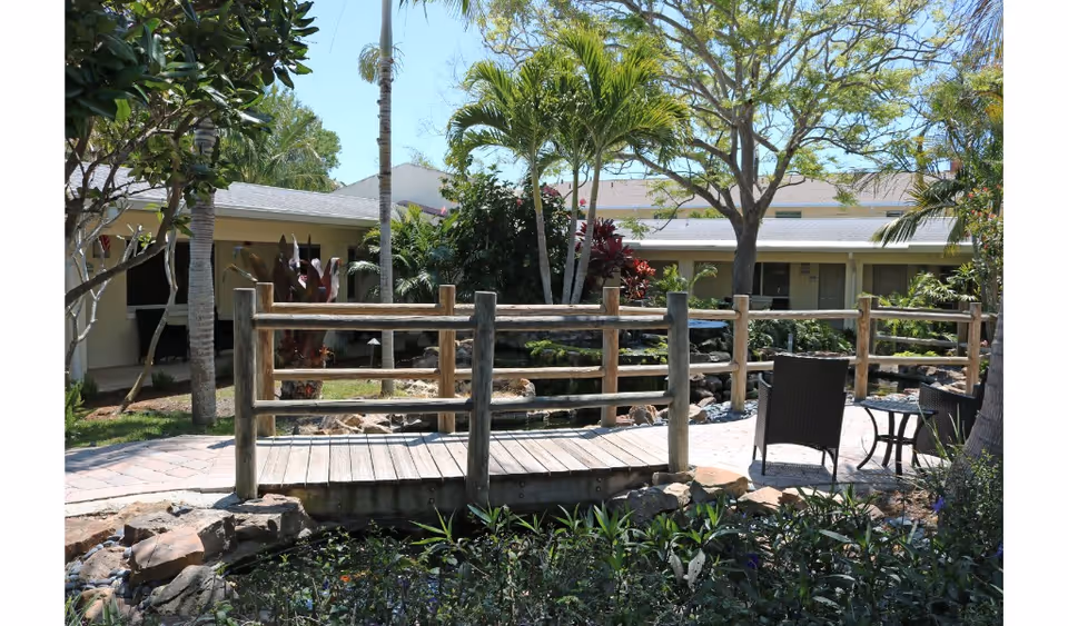 Outdoor garden area at a senior living facility featuring a small wooden bridge over a pond, surrounded by lush greenery, palm trees, and a paved walkway. There are two dark wicker chairs and a small table on the right side of the bridge, with a single-story building in the background under a clear blue sky.