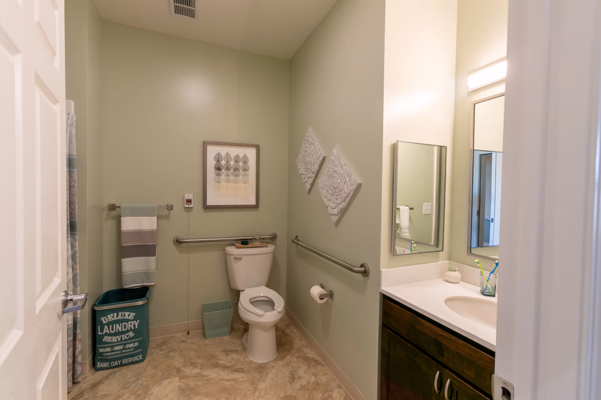 A clean accessible bathroom with a toilet, grab bars, a sink and mirror, towel, and a laundry bin against pale green walls.