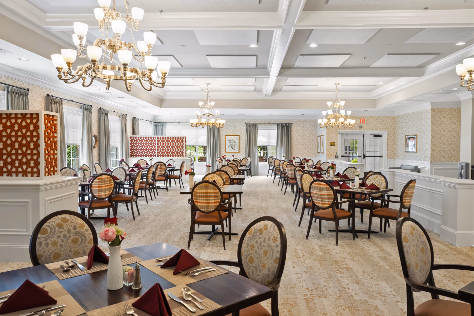 A spacious and elegant dining room with multiple tables set with burgundy napkins, silverware, and small flower vases. The room features large windows with curtains, decorative wooden partitions, and ornate chandeliers hanging from a coffered ceiling. The walls have light-colored wallpaper and white wainscoting, creating a bright and inviting atmosphere.