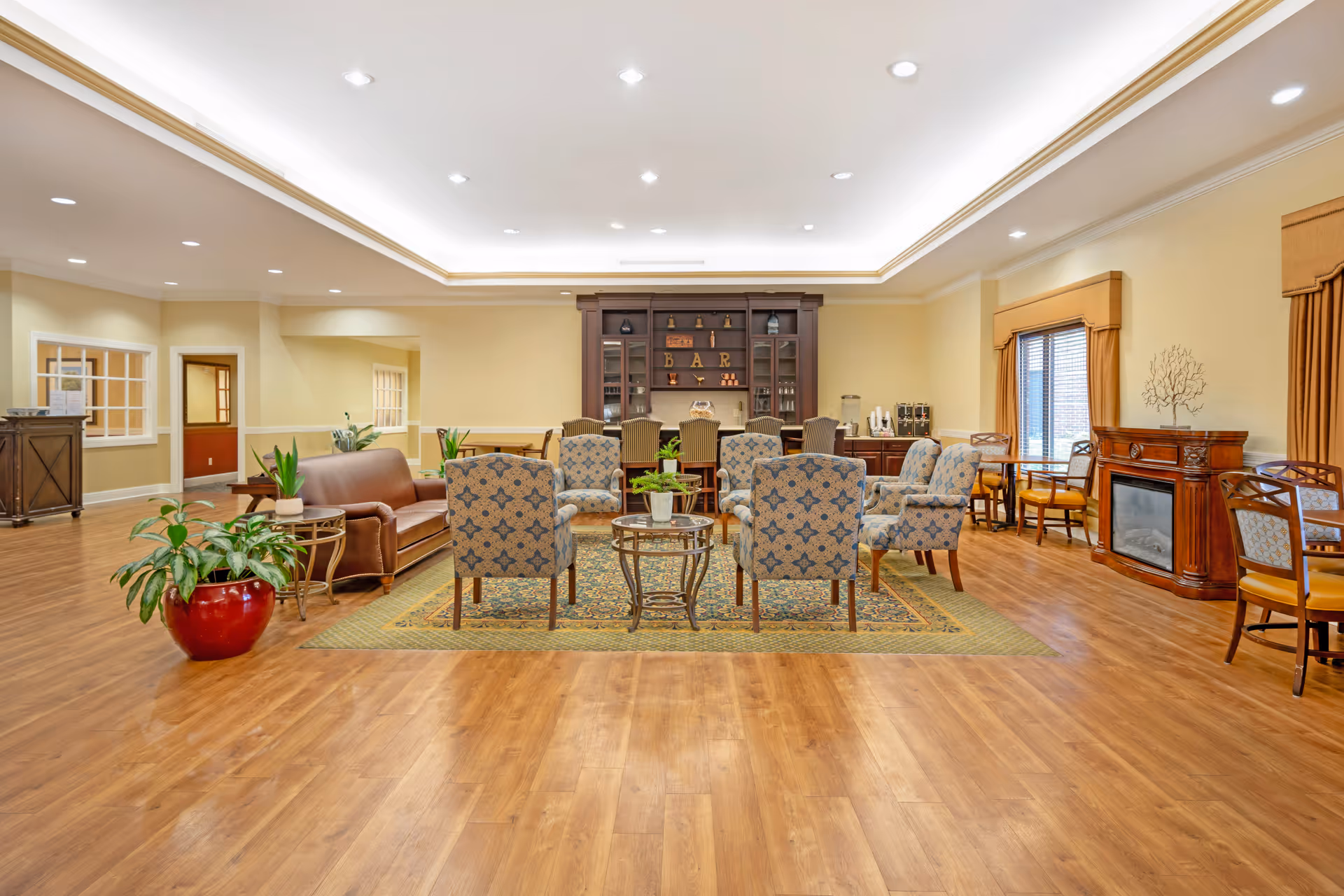 A spacious and well-lit senior living common area with wooden flooring, a seating arrangement of patterned armchairs and a brown leather sofa around a central rug, a wooden cabinet labeled 'BAR' in the background, a fireplace on the right side, and several tables and chairs near windows with curtains.