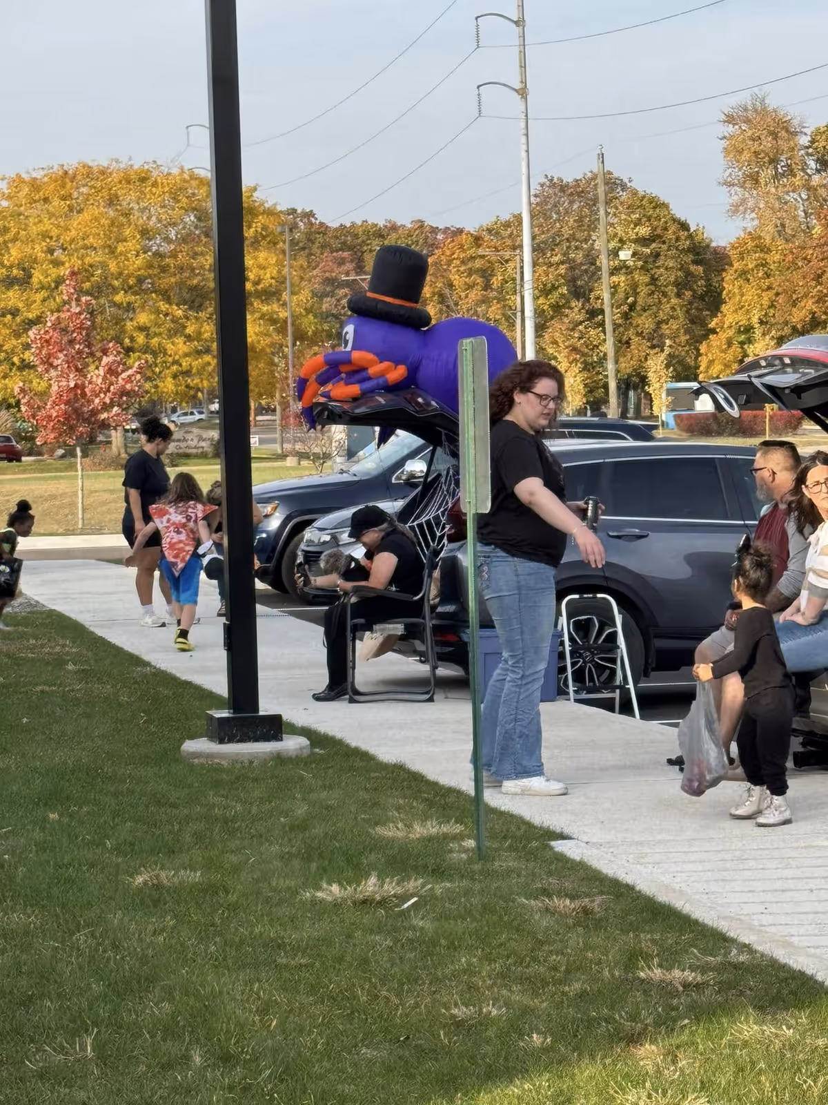 People gathered outside near parked cars on a sidewalk with green grass and trees showing autumn colors in the background. A large inflatable purple spider with a black top hat is attached to the open trunk of a car. Several adults and children are interacting and walking along the sidewalk.