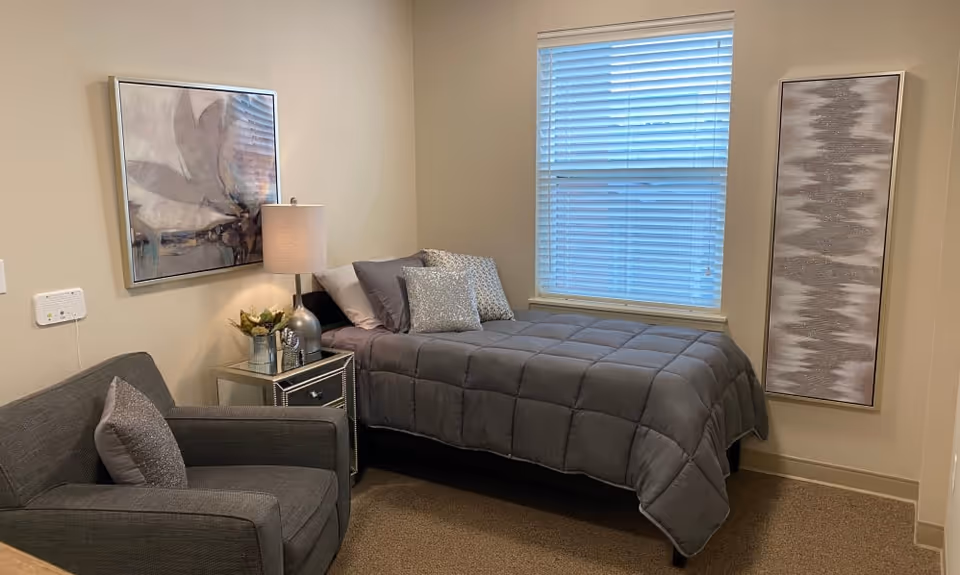 Small furnished bedroom with a single bed, armchair, nightstand and artwork beside a window.