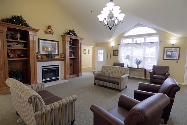 A cozy living room with beige walls and carpeted floor featuring a fireplace flanked by two wooden bookshelves decorated with plants and ornaments. The room has a chandelier hanging from the ceiling, two striped sofas, and three brown leather armchairs arranged around the fireplace. A large window with white curtains lets in natural light, and a small table with a lamp and a vase of flowers is placed in front of the window.