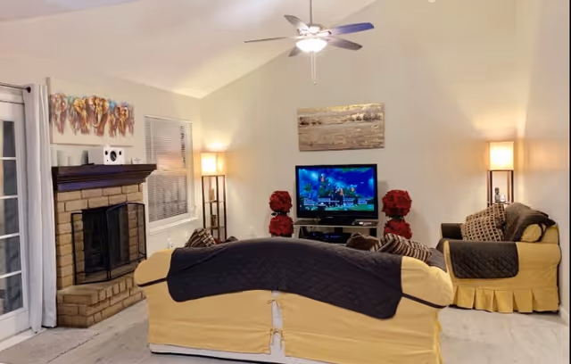 A cozy living room with two yellow sofas covered with dark quilted throws facing a flat-screen TV on a stand. The room features a brick fireplace on the left, two tall floor lamps with warm light, a ceiling fan with light, and decorative artwork on the walls. There are also red floral decorations near the TV stand.
