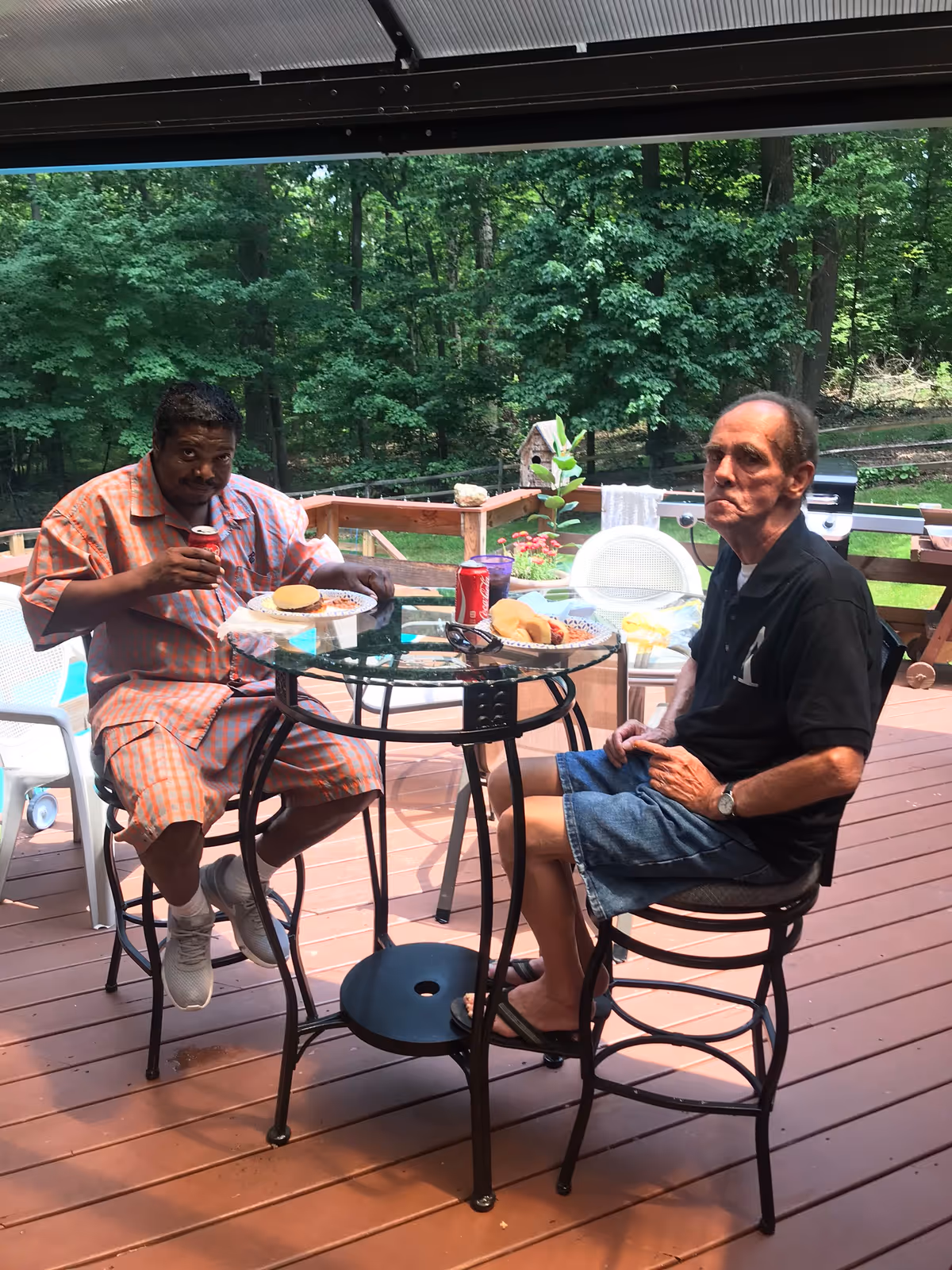 Two men sitting at a round glass table on a wooden deck outdoors, eating a meal with burgers and canned drinks. The background shows a green wooded area with trees and a small birdhouse on the deck railing.