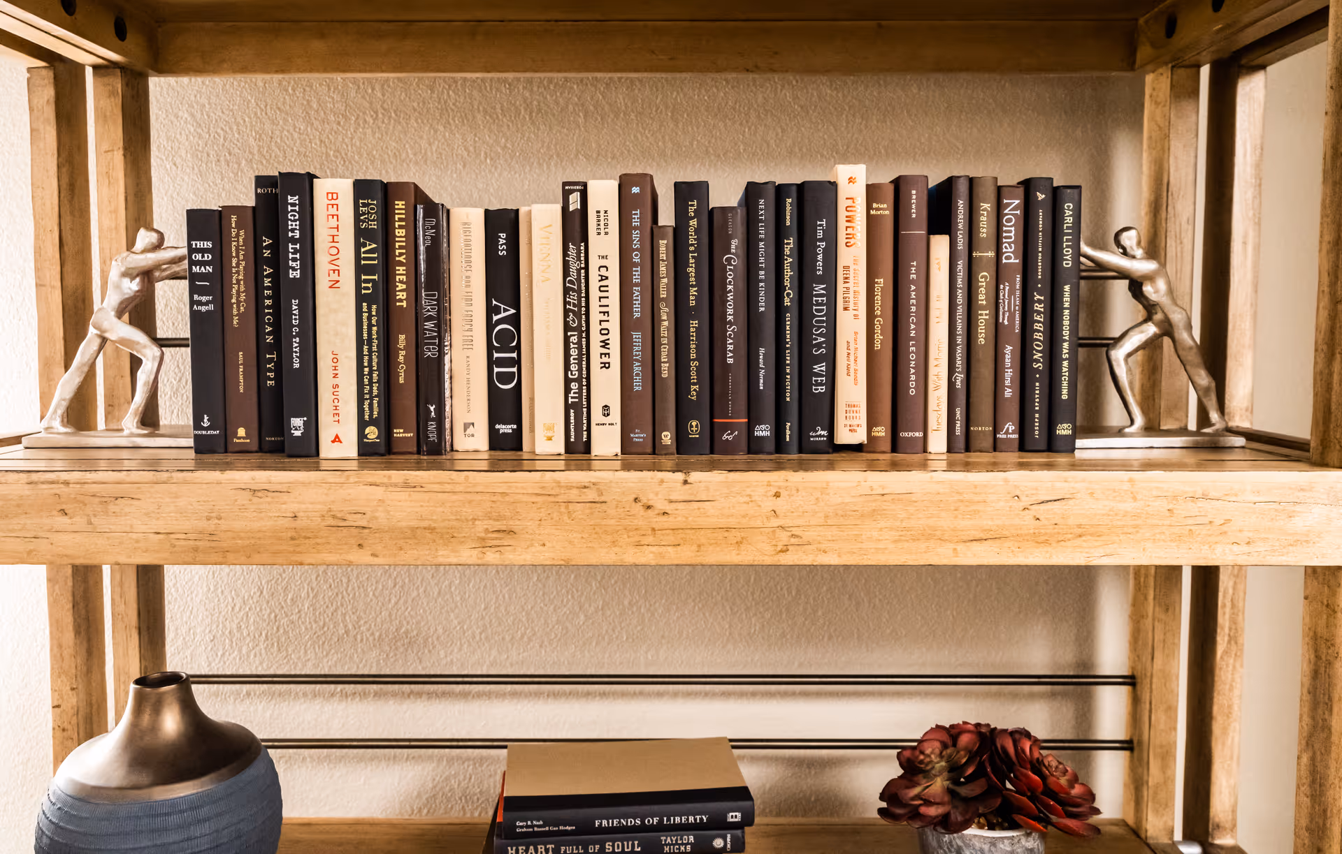 A wooden bookshelf with two metal bookends shaped like human figures pushing against the books. The shelf holds a variety of books with different colored spines. Below the shelf, there is a blue vase and a small potted plant with red flowers.