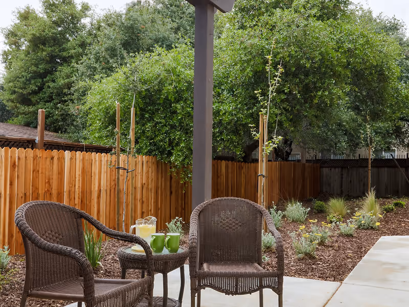 Outdoor patio area with two brown wicker chairs and a small round table holding a pitcher of lemonade and two green mugs. The patio is surrounded by a wooden fence and landscaped garden with small plants and trees.