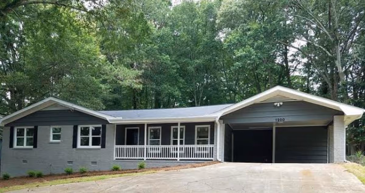 Single-story gray ranch-style house with a covered front porch and attached two-car carport set against a backdrop of trees.