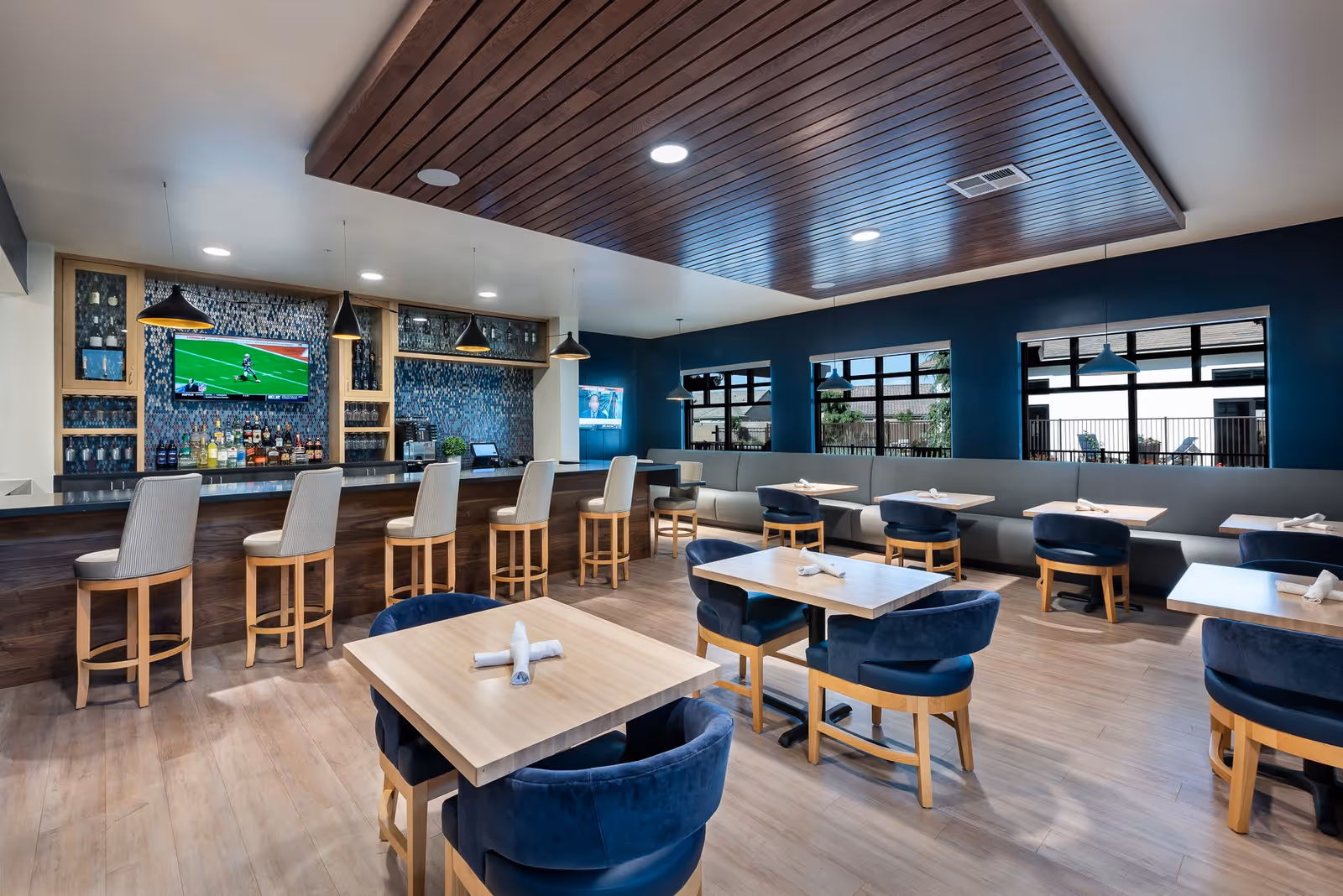 A modern dining area with wooden tables and blue cushioned chairs. There is a bar with high stools and a TV mounted on the wall showing a sports game. The room has large windows, pendant lights, and a wooden ceiling accent.