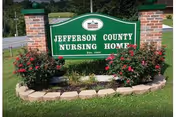 A green sign with white text reading 'Jefferson County Nursing Home Est. 1959' mounted between two brick pillars, surrounded by a flower bed with red flowers and green grass, with trees and a road in the background.