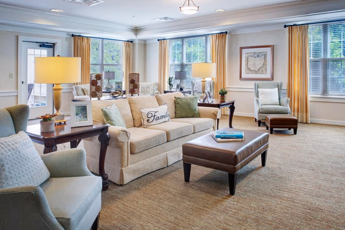 A bright and cozy living room with a beige sofa adorned with green and white pillows, including one with the word 'Family'. There are two armchairs with white cushions, a brown leather ottoman with books on top, and two wooden side tables with lamps and decorative items. Large windows with yellow curtains allow natural light to fill the room, and a framed map hangs on the wall.