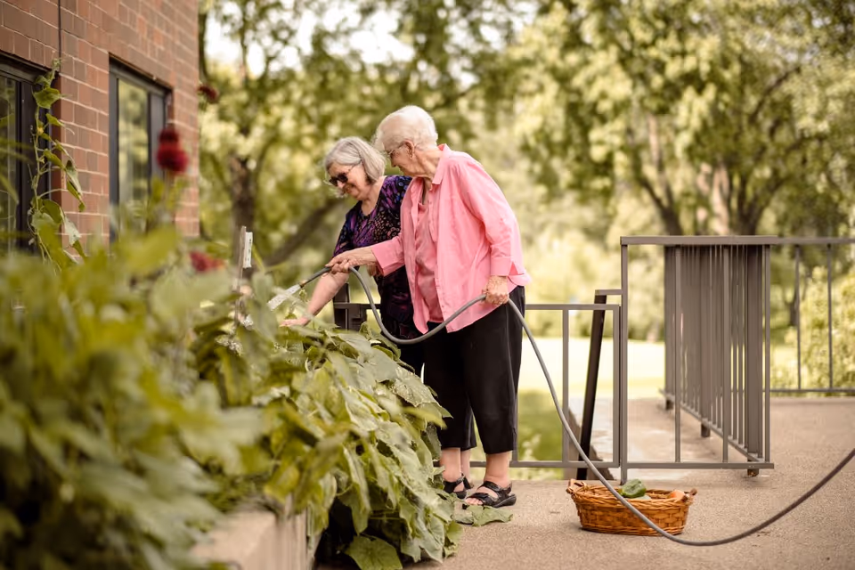 Two elderly women watering plants in a garden area outside a brick building, with one holding a garden hose and the other tending to the plants, surrounded by greenery and trees in the background.