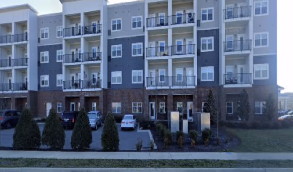 Exterior view of a multi-story residential building with balconies and parked cars in front. The building has a combination of brick and siding on the facade, with several windows and small landscaped areas with bushes.
