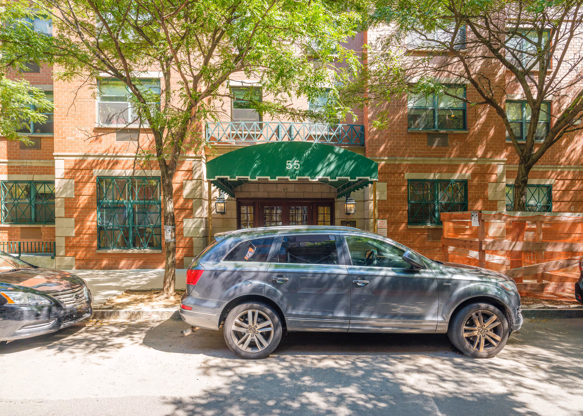 Front entrance of a brick residential building with a green awning marked "55", trees, and cars parked on the street.