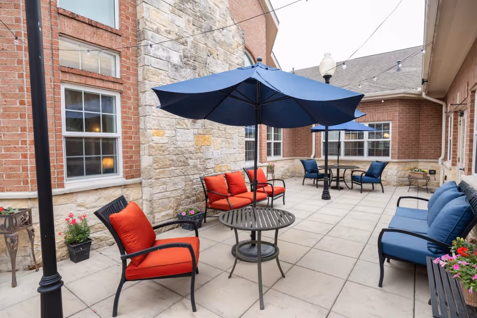 Outdoor patio area with red and blue cushioned chairs, round tables with blue umbrellas, potted flowers, and string lights overhead, surrounded by brick and stone building walls.
