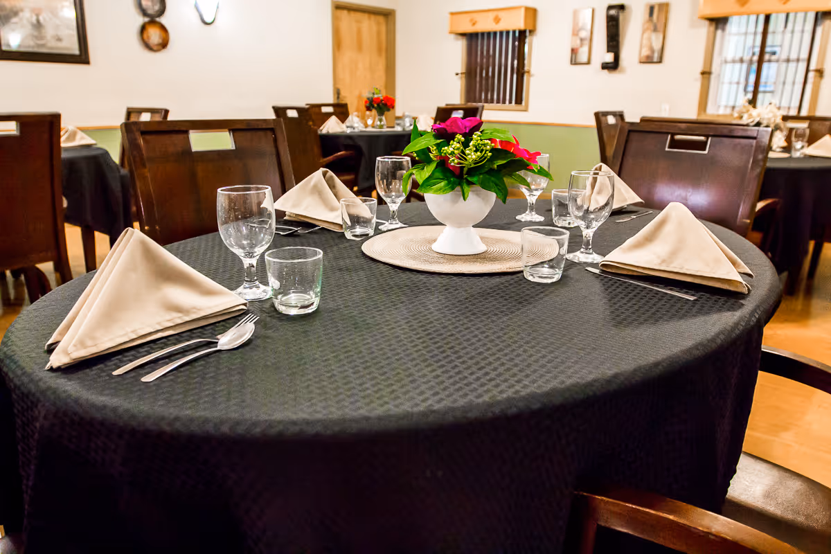 A dining room table set with a black tablecloth, beige folded napkins, clear drinking glasses, silverware, and a white vase with green and red flowers as a centerpiece. The room has wooden chairs, framed pictures on the walls, and a wooden door in the background.