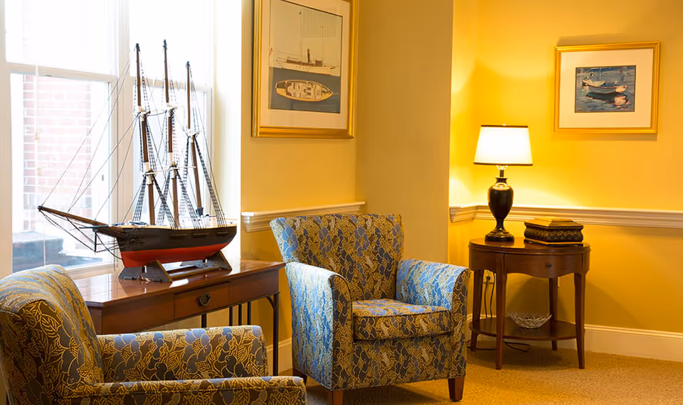 A cozy sitting area with two patterned armchairs in blue and gold, a wooden side table with a model ship on it near a window, a small wooden round table with a lamp and decorative boxes, and framed nautical-themed artwork on yellow walls.