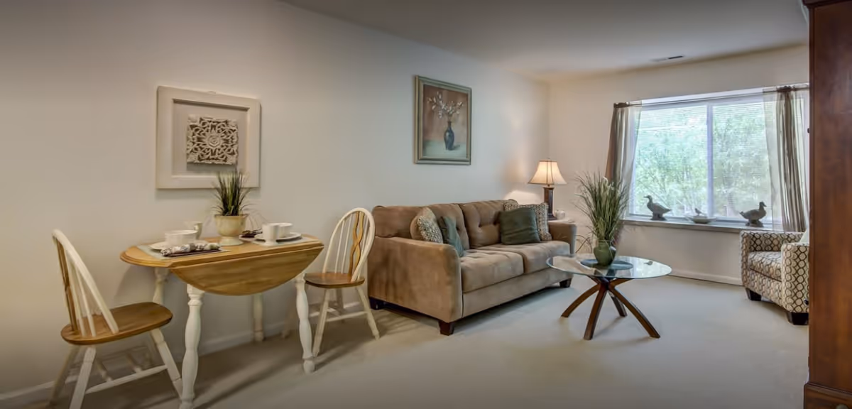 A cozy living room area with a beige sofa adorned with cushions, a glass coffee table with a green vase, a patterned armchair near a large window with brown curtains, and a small wooden dining table with two chairs set with cups and plates. The walls are decorated with framed artwork and a table lamp provides warm lighting.