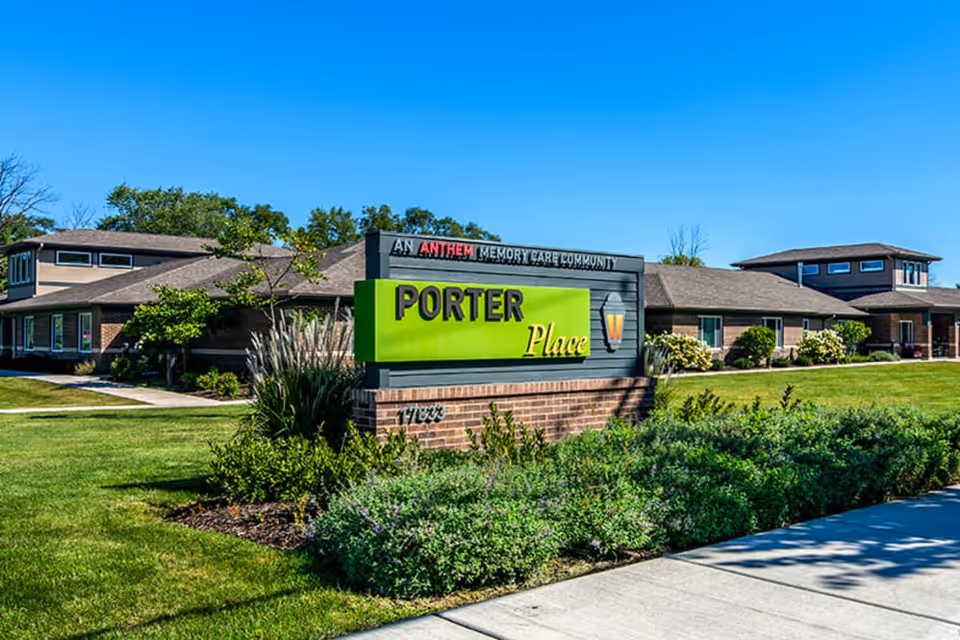 Exterior view of Porter Place, a memory care community building with a large green and black sign in front displaying the facility name. The building is surrounded by well-maintained grass, shrubs, and trees under a clear blue sky.