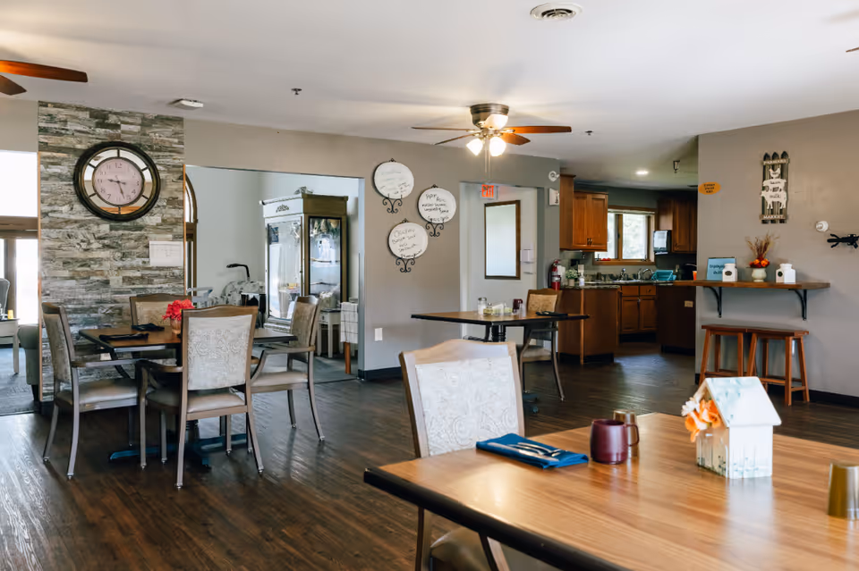 Communal dining area with tables and chairs, a decorative wall clock, and a small kitchen in the background.