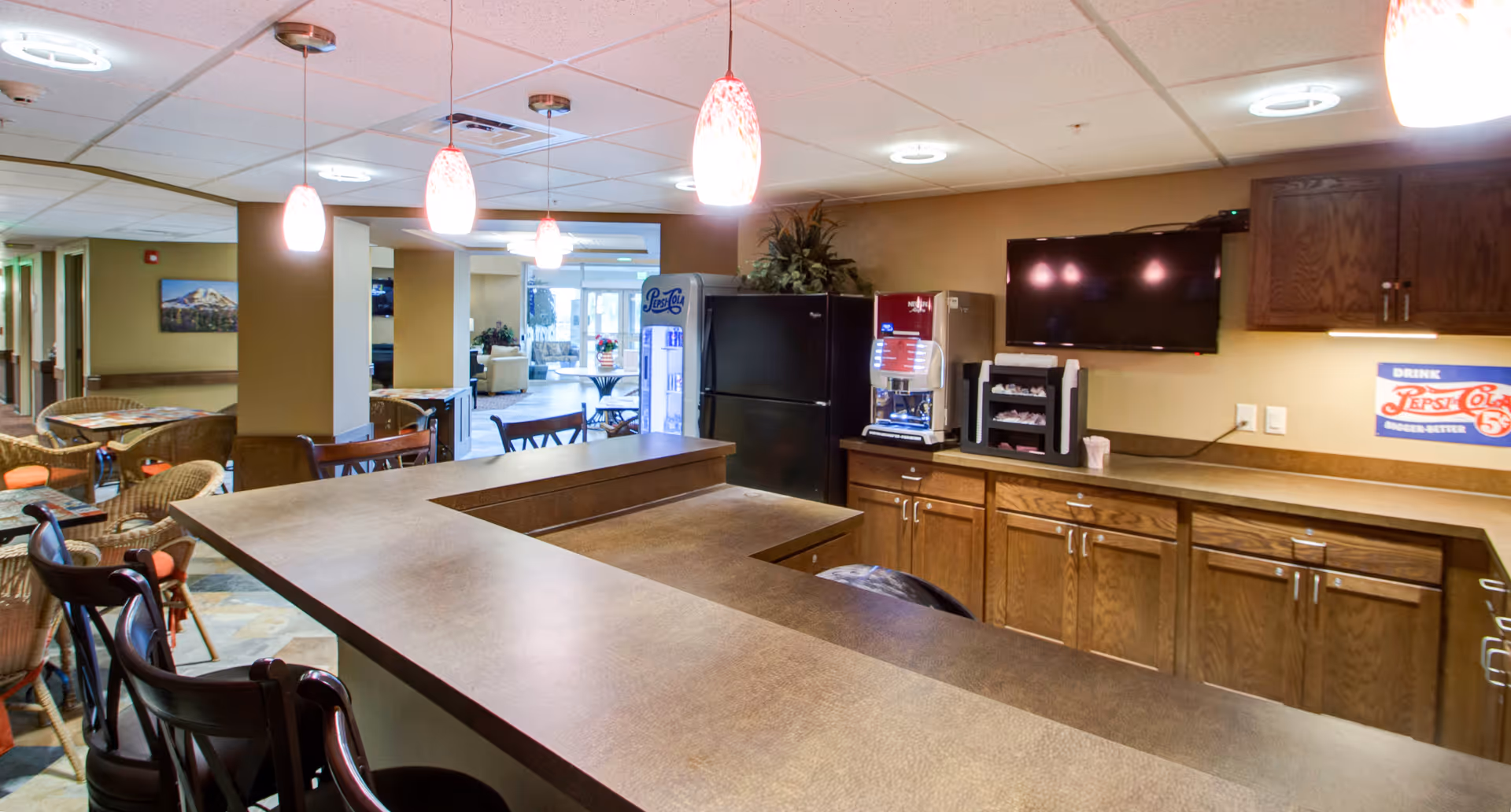 Interior view of a senior living facility common area with a long counter and bar stools in the foreground. Behind the counter are wooden cabinets, a refrigerator, a beverage dispenser, and a wall-mounted TV. The area is well-lit with hanging pendant lights and ceiling lights. In the background, there are tables and chairs arranged for seating, and a hallway leading to other rooms.