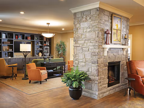 A cozy living room area with a stone fireplace in the center, surrounded by comfortable chairs and a large built-in bookshelf with a TV. There is a green plant in a black pot in front of the fireplace, warm lighting, and wooden flooring.