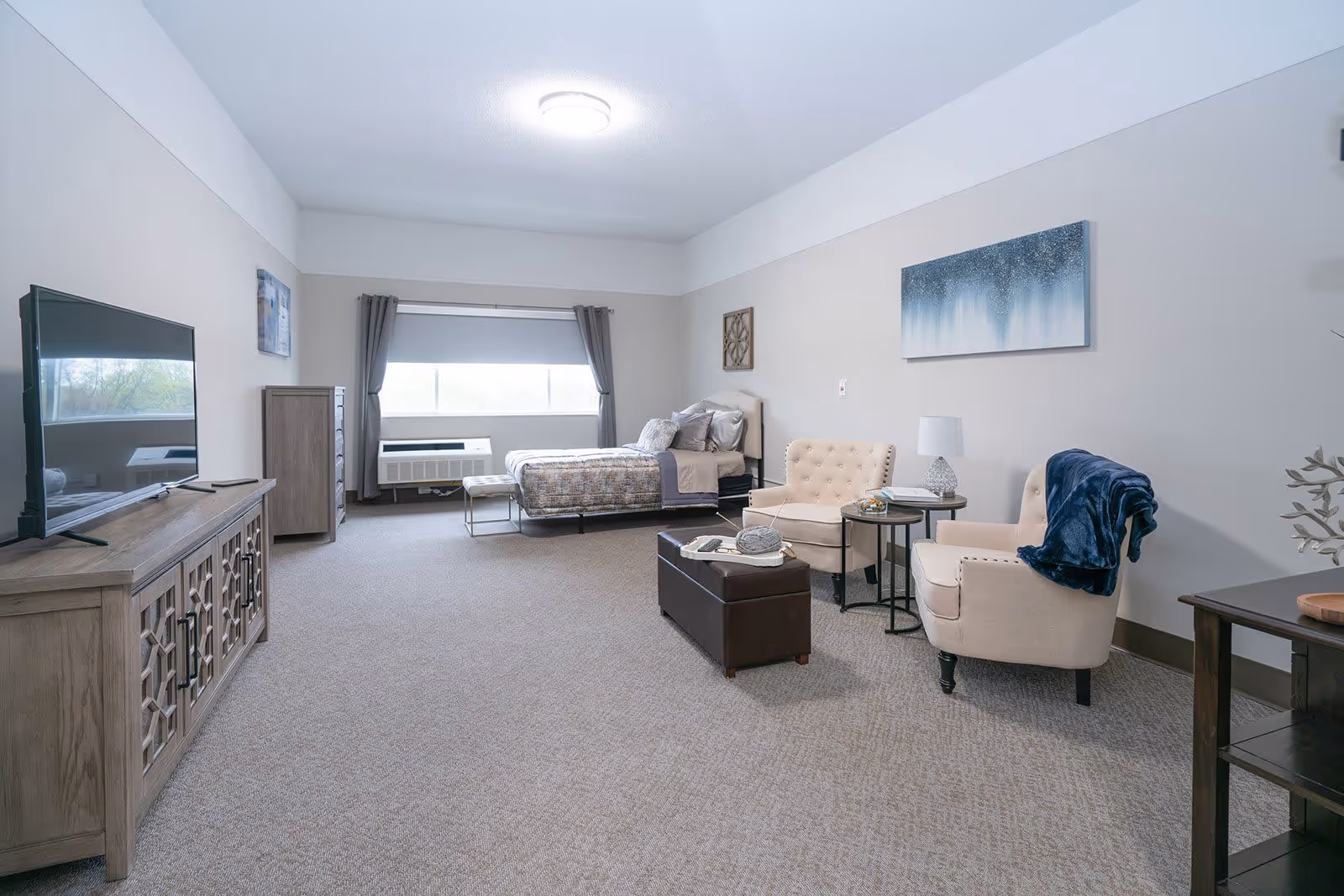 A spacious senior living bedroom featuring a single bed with patterned bedding near a large window with gray curtains. The room includes a wooden dresser with a flat-screen TV on top, two beige armchairs with a dark blue throw blanket on one, a small round side table with a lamp, and a dark brown ottoman in the center. The walls are light-colored with modern artwork hanging above the chairs.