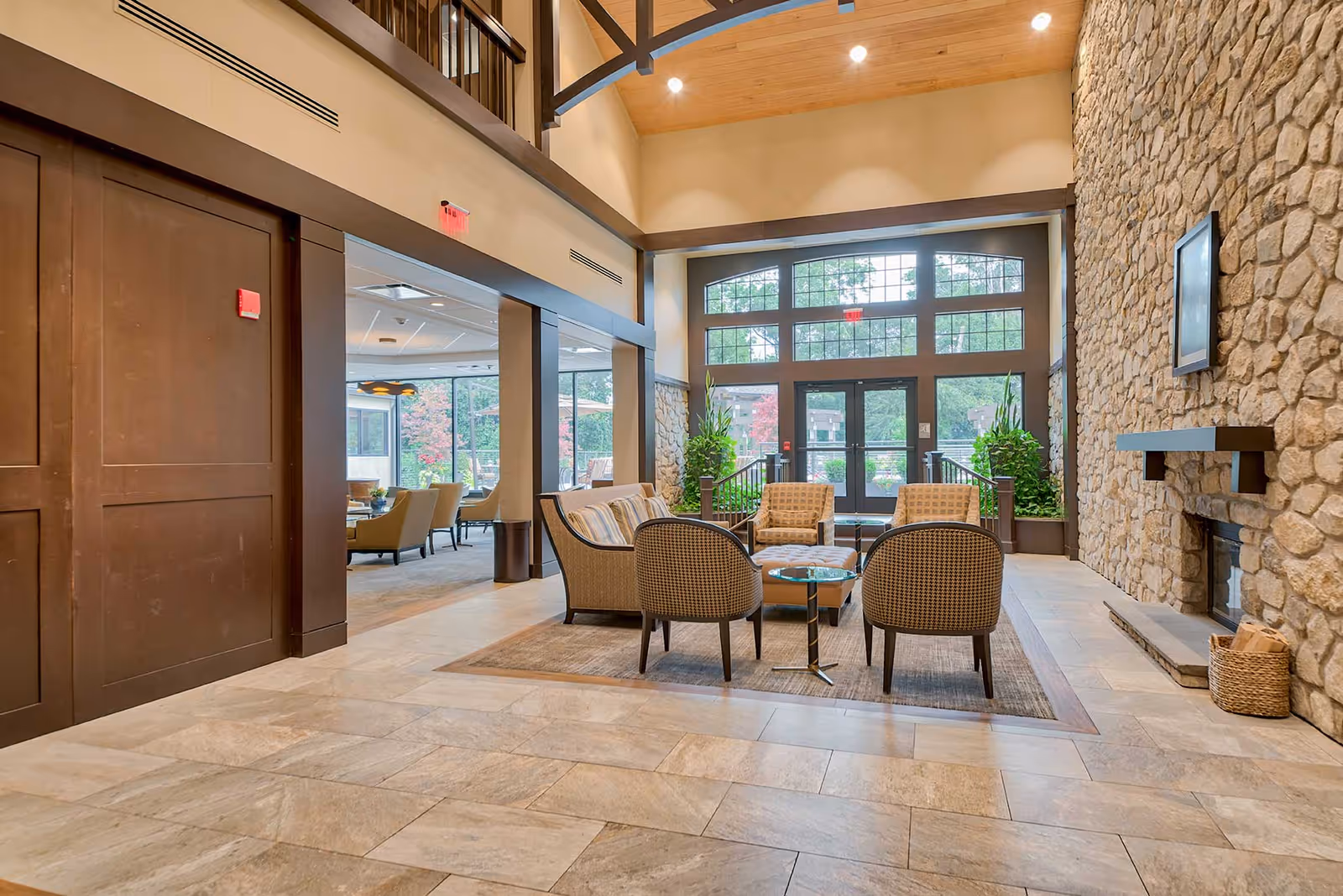 A spacious and well-lit common area in a senior living facility featuring a seating arrangement with four chairs and a small round table on a rug. The room has a high ceiling with wooden beams, large windows and glass doors letting in natural light, stone walls with a fireplace, and green plants near the entrance.