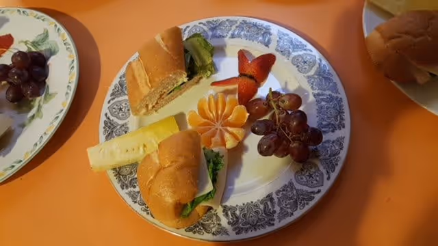 A plate with two small sandwiches, a peeled and segmented orange, a strawberry cut to resemble a flower, and a small bunch of red grapes on an orange table. Another plate with grapes is partially visible on the left, and a sandwich on a bun is partially visible on the right.