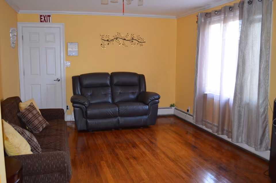A cozy living room with yellow walls and polished wooden floors. The room features a dark brown leather loveseat and a brown fabric sofa with patterned and yellow cushions. A decorative metal wall art piece hangs above the loveseat. A window with sheer gray curtains allows natural light to brighten the space. A white door with an exit sign above it is visible on the left side of the room.