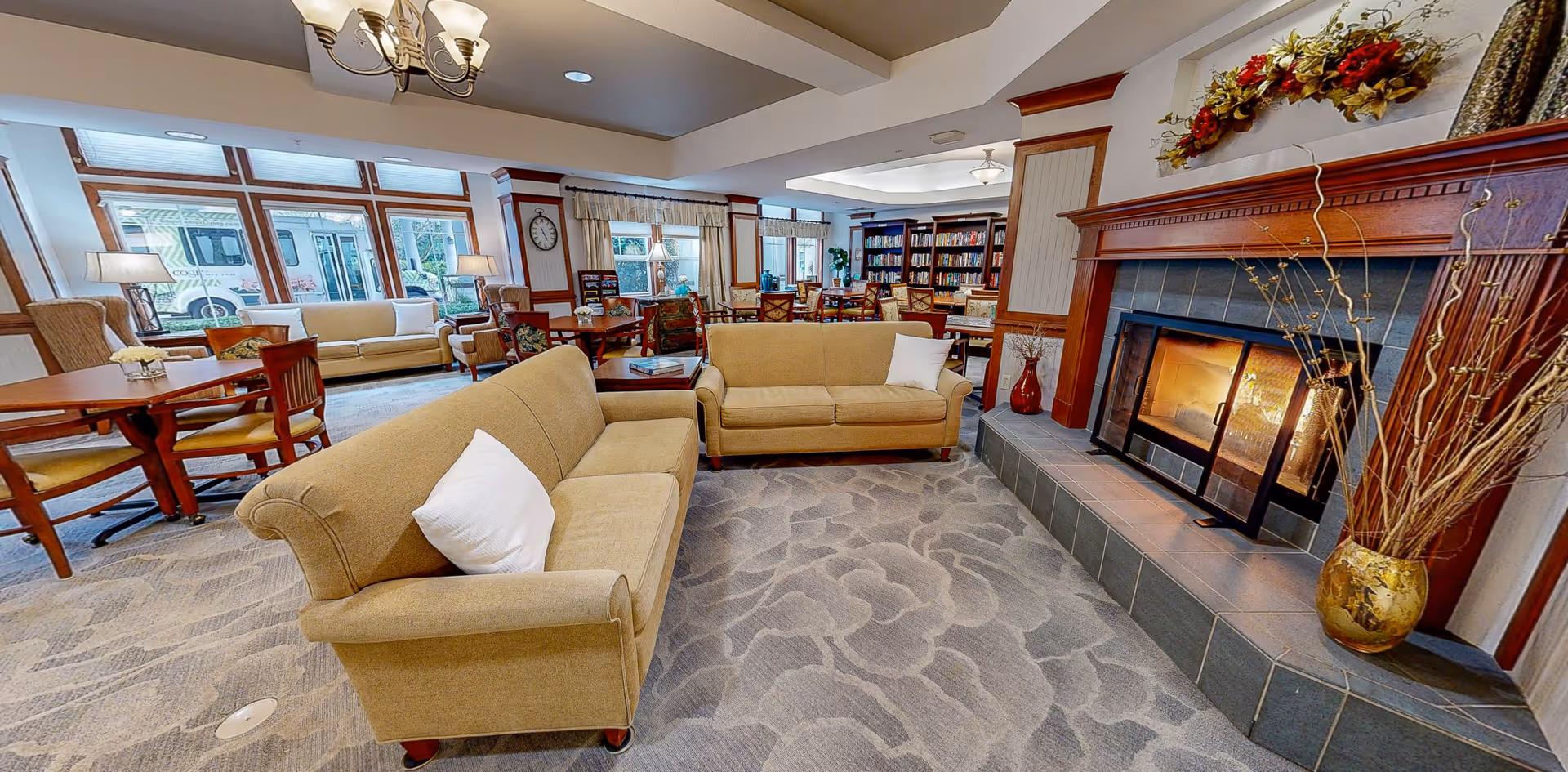 A cozy senior living facility common area with beige sofas, a lit fireplace with a wooden mantle decorated with flowers, several wooden tables and chairs, large windows letting in natural light, and a bookshelf filled with books in the background.
