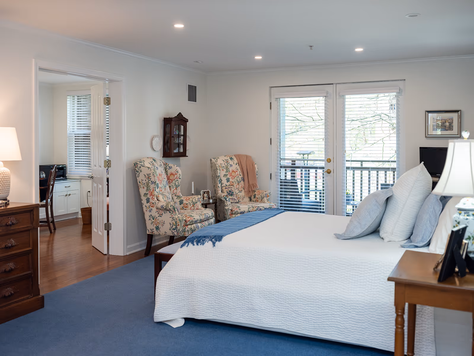 A bright and cozy bedroom in a senior living community featuring a neatly made bed with white bedding and multiple pillows. Two floral upholstered armchairs are placed near a glass door leading to a balcony. A wooden dresser with a lamp and a side table with a lamp and framed photos are also visible. The room has light-colored walls, blue carpet, and an adjacent room with a desk and chair can be seen through an open door.