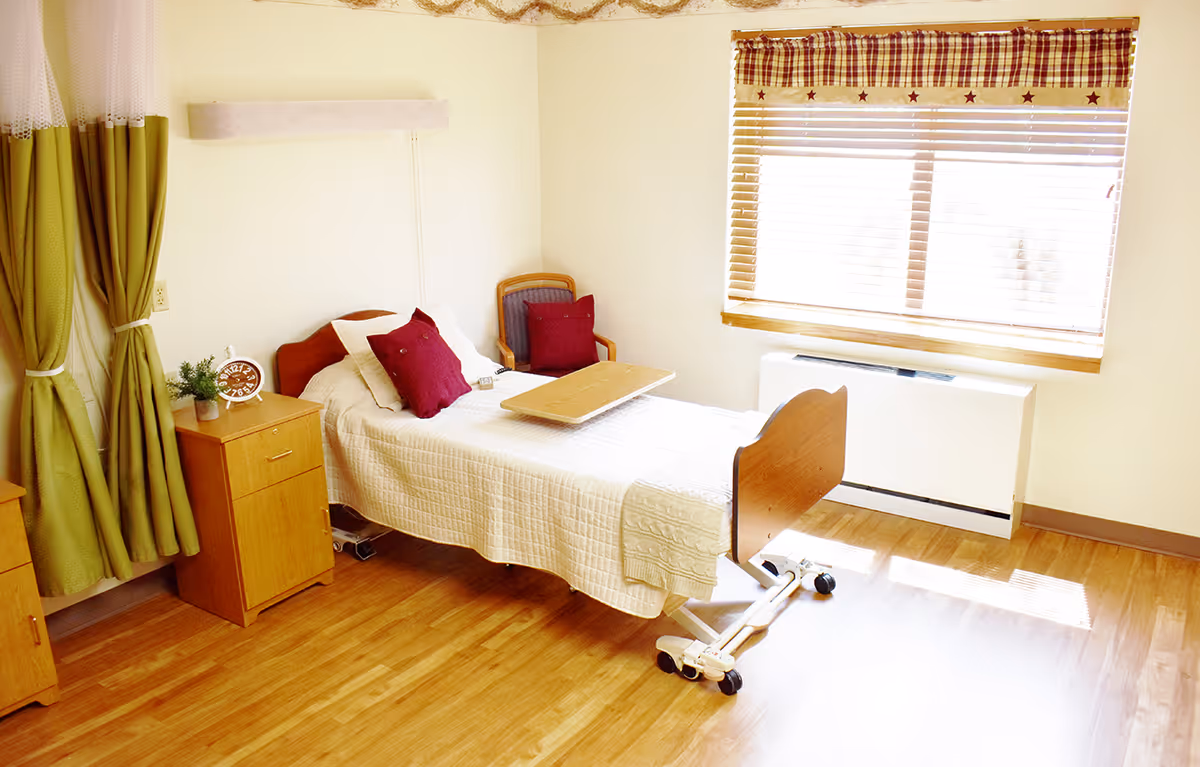 A bright and clean senior care bedroom with a single hospital-style bed covered in a white quilt, two red pillows, a wooden overbed table, a wooden nightstand with a clock and plant, a wooden chair with a red cushion, green curtains, and a window with blinds and a plaid valance.