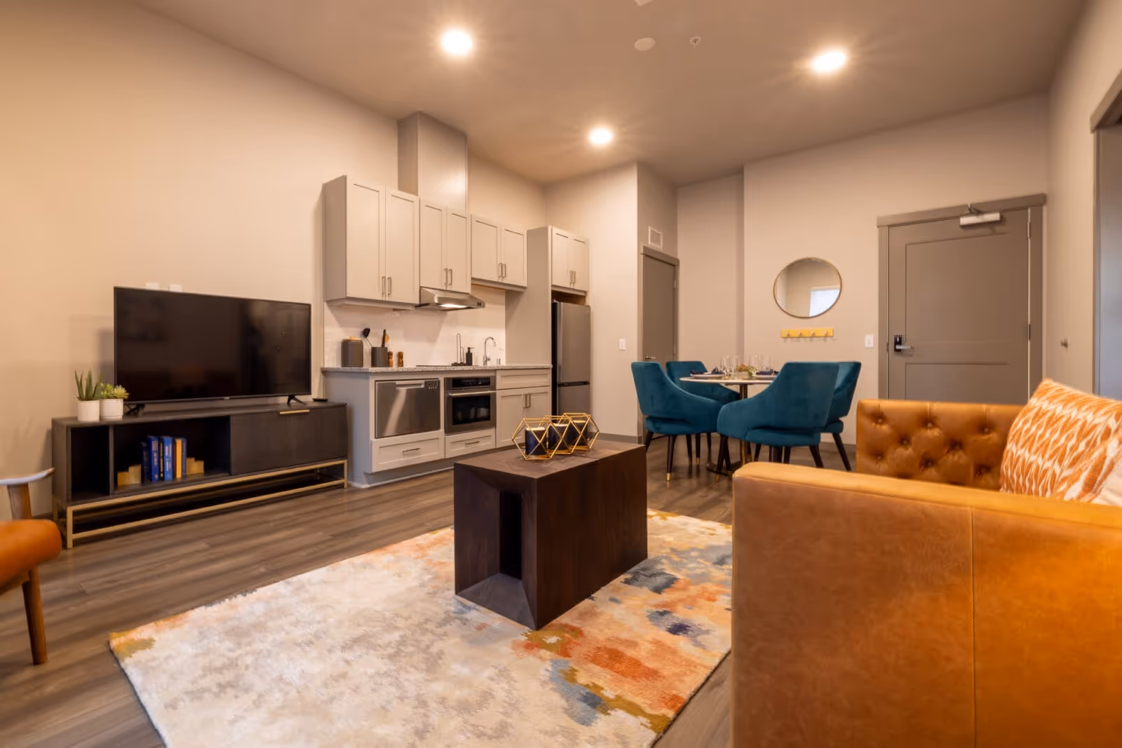A modern living area in an assisted living facility featuring a tan leather tufted sofa with a patterned pillow, a dark wooden coffee table on a multicolored rug, a flat-screen TV on a black media console with books and plants, and a compact kitchen with gray cabinets, stainless steel appliances, and a dining table with four teal upholstered chairs.