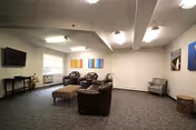 A spacious living room area with a brown leather sofa, two matching armchairs, and a gray armchair. There is a large ottoman in the center, a flat-screen TV mounted on the wall, and colorful abstract paintings hanging on the walls. The room has carpeted flooring and white walls with ceiling lights.